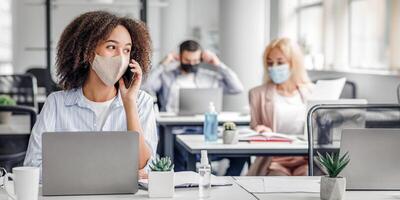 Working with clients remotely after returning from quarantine. African american woman in protective mask speaks on phone, sitting at table with laptop in modern office interior with colleagues, empty space photo