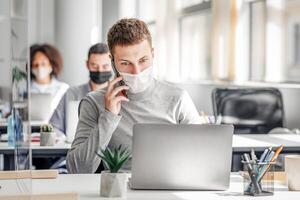 Social distance and online work with client remotely. Busy man in protective mask talking on smart phone sitting at office in workplace with laptop and glass board during coronavirus, copy space photo