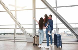 Weekend Travel. Family Of Three Standing Near Window In Airport And Pointing Away, Rear View Of Young Parents And Their Little Daughter With Suitcases Waiting For Flight At Terminal, Copy Space photo
