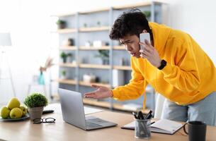 Confused Discontented Asian Man Talking On Phone Using Laptop Having Problem With Computer Or Internet Connection At Home. Displeased Male Customer Calling To Hotline Service And Complaining photo