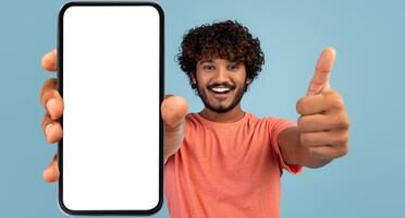 Handsome indian millennial guy showing brand new smartphone with empty white screen and thumb up, smiling at camera over blue studio background, mockup, mobile application concept, panorama photo