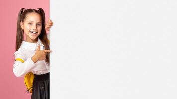 Pupil going to school. Little girl in school uniform peeps out of billboard with empty space and points her finger at it, studio shot photo