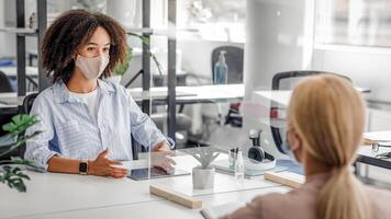 Interview. Young business workers from different ethnicities in protective mask talking to each other in office through partition to prevent COVID-19 infection photo