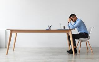 Profile side view of young Middle Eastern man suffering from headache at workplace, having problems thinking about troubles. Stressed guy in glasses sitting at desk using computer, free copy space photo