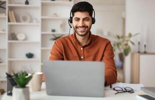 Portrait Of Smiling Middle Eastern Male Manager In Headset Working On Laptop Computer In Modern Home Office, Happy Guy Sitting At Desk And Consulting Clients Online Using Pc, Free Copy Space photo