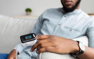 Unrecognizable Black Man With Pulse Oximeter On Hand Measuring Oxygen Saturation Level At Home. Pulseoxymeter Medical Device, Pulseoxymetry Clip Machine Monitorin Ox Rate. Cropped, Selective Focus photo