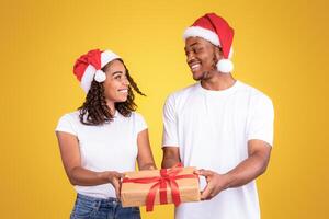 African American Couple Offering Christmas Gift To Camera, Holding Wrapped Xmas Present Box Posing On Yellow Studio Background, Wearing Santa Hats. Winter Holidays Celebration Concept photo