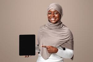 Religious african muslim lady in hijab holding and pointing at digital tablet with black screen, demonstrating free place for new app or website mockup design, standing over light background in studio photo