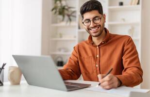 Portrait of young smiling Arabic man in glasses using laptop sitting at desk, writing in notebook. Happy guy working remotely, watching webinar training studying online, taking notes signing documents photo