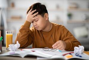 Exhautsed chubby asian kid teen boy doing homework alone, sitting at table full of exercise books, notepads, glass of orange juice, touching his head, experiencing difficulties with studying photo