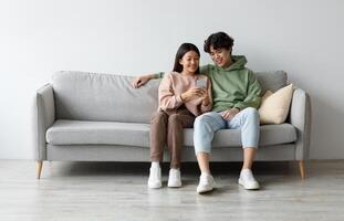 Happy young Asian couple using cellphone, browsing internet, chatting online, relaxing on sofa at home, free space. Cheery millennial family checking new mobile application, resting on couch together photo