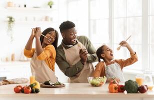 Positive Black Family Of Three Cooking And Having Fun Singing Favorite Song Holding Spoons Preparing Dinner Together Standing In Modern Kitchen Indoor. Weekend Leisure, Healthy Food Recipes Concept photo