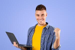 Online travel agency. Positive young guy with laptop booking tickets on web and making YES gesture over blue studio background. Millennial man making hotel reservations for vacation through internet photo