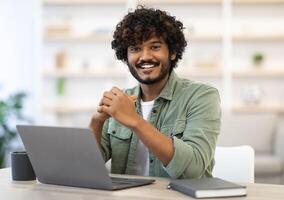 Cheerful attractive indian guy in casual outfit programmer posing at workdesk at office, eastern freelancer sitting in front of computer at home, working online, smiling at camera, copy space photo