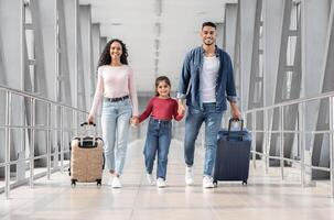 Ready For Vacation. Happy Middle Eastern Family Of Three Walking With Suitcases At Airport Terminal, Smiling Arab Parents With Little Daughter Holding Hands, Enjoying Travelling Together, Copy Space photo