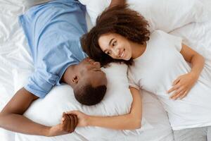 Happiness together at home, rest and relax at weekend at covid-19. Smiling millennial african american woman and man lie on soft white bed and look at each other in bedroom, top view, empty space photo