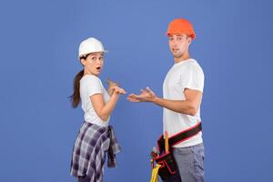 Concerned couple of workers in protective helmets shrugging, making i dont know gesture. Surprised man and woman having problems during house renovation, standing on blue studio background, copy space photo