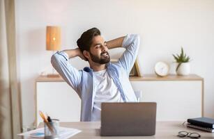 Relaxed Eastern Man Leaning Back In Chair, Resting After Online Work With Laptop, Millennial Arab Guy Sitting At Table With Computer In Home Office With Hands Behind Head And Looking Aside, Free Space photo