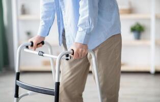 Cropped view of elderly man walking with frame at home, closeup. Unrecognizable senior male using medical equipment to move around his house. Disabled older person in need of professional help photo