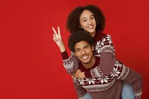 Have fun together during Christmas and New Year holidays. Cheerful young african american male holds female on back, lady shows peas sing with hand and watch at camera, isolated on red background photo