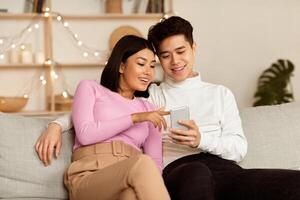 Chinese Millennial Couple Using Smartphone Relaxing On Weekend Sitting On Couch At Home, Spending Evening Together. Mobile Application, Phone Users Browsing Internet, Gadget Lifestyle Concept photo