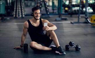 Fitness Application. Smiling Middle Eastern Sportsman Using Smartphone At Gym While Relaxing After Training, Handsome Arab Male Athlete Browsing New Mobile App With Workout Tutorials, Copy Space photo