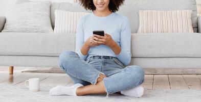 Social networks, app, blog and surfing in internet during self-isolation of covid-19. Smiling young african american lady sits on floor in lotus position with cup of tea and typing in phone at home photo