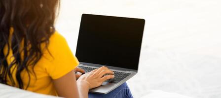 Unrecognizable black lady typing on laptop with blank screen on comfy couch at home, mockup for design. Young female freelancer working online, student making project, space for your website photo