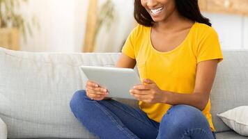 Full length portrait of happy black woman using tablet computer on couch in living room. African American lady with touch pad browsing web, watching or communicating on webcam photo