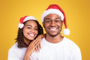 Christmas Portrait Of Joyful Black Couple In Santa Hats Hugging Posing Over Yellow Studio Background, Smiling To Camera. Xmas, New Year And Winter Holidays Celebration. Panorama photo