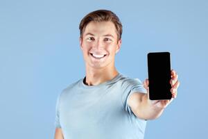 Happy young sportsman showing smartphone with empty screen over lilac studio background, mockup for mobile app design. Strong millennial guy holding cellphone with space for website template photo