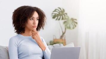 Solving problem, creating idea, work from home and taking care of health. Thoughtful millennial african american woman sits on sofa with laptop in living room interior and thinking about startup photo