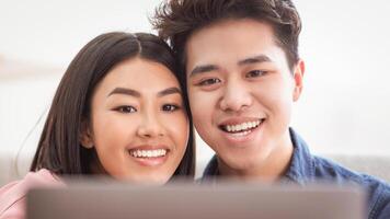 Chinese Couple At Laptop Watching Movie Online Sitting On Sofa At Home, Relaxing Together On Weekend. Happy Family Using Computer Browsing Internet, Modern Lifestyle Concept. Selective focus photo