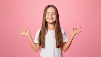 Calming, meditation, mood. Little child with closed eyes practices yoga, isolated on pink background, studio shoot photo