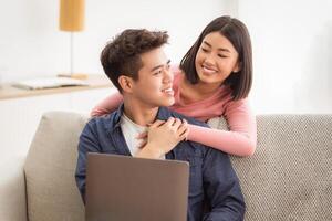 Asian Married Couple Using Laptop Computer Browsing Internet Together Sitting On Sofa In Living Room At Home. Korean Family Enjoying Weekend. Empty Space For Text photo