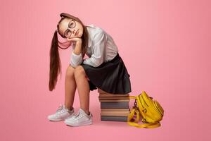Tired pupil dreams. Schoolgirl in glasses with ponytails and backpack sits on stack of books and looks at empty space, on pink background, studio shoot photo