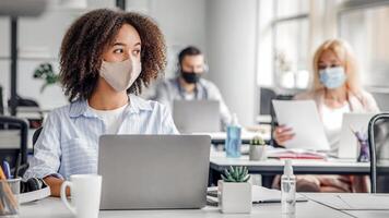 Creating an idea at work brainstorming for project. Serious african american lady in protective mask looks at window working at computer in modern office during outbreak of coronavirus, copy space photo