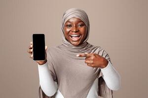 Cheerful Black Islamic Lady In Hijab Pointing At Smartphone With Black Blank Screen, Demonstrating Modern Gadget With Copy Space For Your Design, Standing Over Light Studio Background, Mockup Image photo