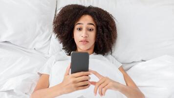 Amazing or bad news, strange offer during covid-19 lockdown. Shocked young zoomer african american female lies in soft bed and looks at smartphone, reading message in bedroom, top view, free space photo