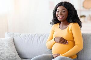 Joyful black pregnant woman hugging her big tummy, communicating with her baby, sitting on sofa at living room. Happy african american expecting lady spending time at home, looking at copy space photo