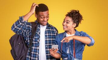 Black Woman Recommending Travel Application Showing Phone To Confused Friend Preparing For Trip Standing On Yellow Background In Studio. Mobile App And Service For Tourists Concept photo