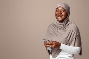 Mobile Communication. Cheerful Religious Black Woman In Hijab Posing With Smartphone In Hands, Holding Cellphone And Smiling At Camera, Enjoying New App, Standing Over Light Background In Studio photo