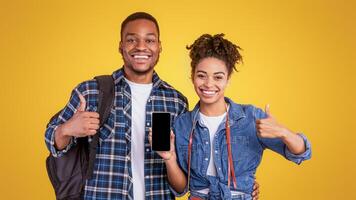 Happy African Travellers Couple Showing Smartphone Empty Screen Gesturing Thumbs Up, Recommending Mobile Travel App Standing On Yellow Studio Background. Approving Application For Tourists Concept photo