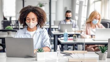 Modern devices for working in coworking office during virus outbreak. African american woman in protective mask typing on laptop at table with smartphone, antiseptic and protective glass, empty space photo