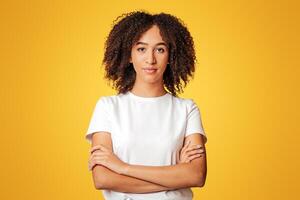 Smart student and human emotions. Serious confident young african american woman with curly hair in white t-shirt with crossed arms, isolated on yellow background, empty space, studio shot photo
