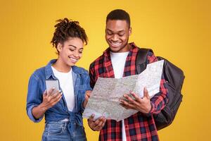 Travel App. Happy Black Tourists Couple With Smartphone And Map Choosing Destination For Vacation Standing Over Yellow Background In Studio photo