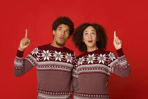 Amazing offer for gifts for New Year and Christmas. Surprised african american couple in sweaters pointing up and looking at free space, woman with open mouth isolated on red background, studio shot photo