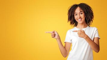 Advertisement and presentation concept. Smiling joyful millennial african american lady with curly hair in white t-shirt pointing with fingers to empty space isolated on yellow background, studio shot photo