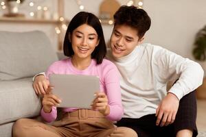 Chinese Spouses Watching Movie Online Via Digital Tablet Computer Sitting Of Floor At Home. Weekend Relaxation, Online Movie Player Application Concept. photo