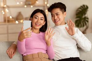 Japanese Couple Making Call Using Mobile Phone Waving Hand To Smartphone Sitting On Sofa At Home. Distant Communication, Online Conference Application For Cellphone Concept photo